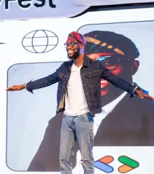 A speaker in a denim jacket and patterned headwrap, arms open, presenting to the room.